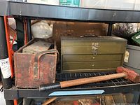 Photo showing wooden tool carrier, green metal toolbox with drawers, rusted small metal toolbox, and a red sledgehammer with wooden handle lying on a shelf.