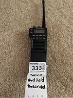 Full view of Standard Communications marine handheld transceiver with antenna, placed vertically on carpet with label noting 'marine handheld transceiver'.