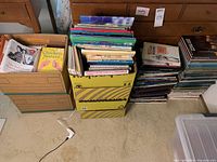 Photo of several boxes filled with various books including fiction, biography, travel, religious, and children's books. Books are stacked both inside and beside boxes on carpeted floor.