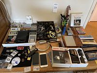 Full overview of the desk supplies lot showing calculators, pens, lapel pins, iPhones, magnifying glass, boxed microcassette recorders, and lighting accessory.