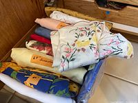 Assortment of folded and rolled linens including round tablecloths labeled with 68" diameter and various colors and patterns; fabric appears lightly used