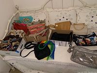 Overview of assorted purses and bags on a bed showing various colors, sizes, and styles including a silver crossbody and woven basket bags.