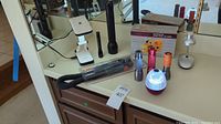 Photo showing assorted small lights including a black tactical flashlight, white foldable task light with phone holder, red spherical LED light, silver and orange flashlights, and packaged flexible hose light attachment on countertop.