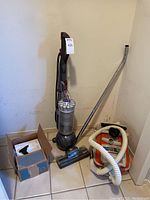 Photo showing Dyson DC40 upright vacuum next to orange and white Hoover canister vacuum and cardboard box of accessories in corner.