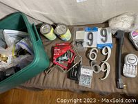 Overview of assorted hardware on a couch showing new lock box, packaged house numbers, a red tool clamp, paint cans, screws pack, and a hammer.