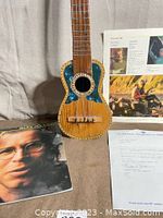 Full view of charango instrument leaning against backdrop with album cover and letter visible behind