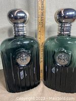Front view of two dark green glass display bottles with silver-tone metallic caps and embossed medallions, a ruler behind them shows height.