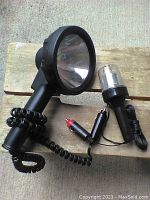 Four auto lights shown together: two large spotlights with coiled cords and vehicle power plugs, one smaller flood light, and one metal-caged trouble light, all placed on wooden surface.