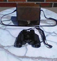Front view of brown carrying case with strap and Bushnell binoculars placed on white surface.