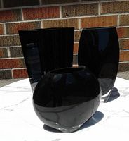 Three black art glass vases arranged on a white marble surface, showing their shapes and clear bases against a brick wall background