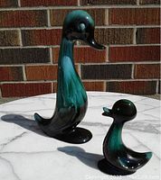 Front and side view of two ceramic duck figurines with teal and black glaze on a marble tabletop against a brick background.