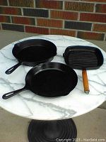 Photo of three cast iron pans on white marble top table against brick background showing entire lot.