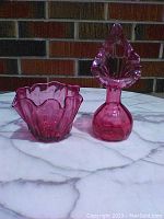Overview of both cranberry glass items, bowl and vase side-by-side on white marble surface against brick background.