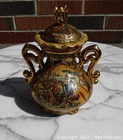 Front view of lidded urn jar showing painted scene of people and elaborate handles with gold accents.