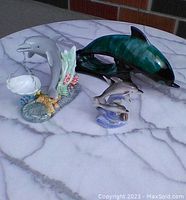 Set of three dolphin figurines displayed on white marble table: large Blue Mountain Pottery dolphin, gray dolphin with coral holding dish, pair of small dolphins on wave base.