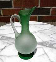 Green glass pitcher displaying gradient from frosted light green to dark green with curved handle and spout on marble surface with brick background.