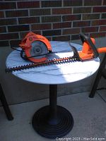 Orange Black & Decker Skilsaw and hedge trimmer on a round table outside, side view