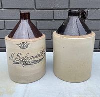 Front view of two antique stoneware jugs side by side, showing advertisement on one jug and plain surface on the other.