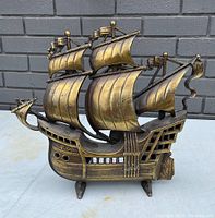 Front angle photo showing the detailed cast iron sailing ship with bronze/gold finish, sails and flags, set against a gray brick background.
