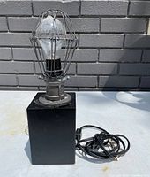 Front and side view of industrial desk lamp with metal cage protecting bulb, black square base, and visible cord.