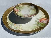 Two-tiered porcelain serving plate with hand-painted roses and heavy gold ribbing on rims, shown outdoors on a light surface.