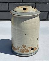 Side view of cylindrical pottery piece showing ventilation holes and floral pattern around base under natural light.