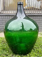 Front view of a large green glass demijohn placed outdoors, showing the wide body and narrow neck with smooth glass surface.