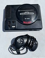 Top view of Sega Genesis Model 1 console showing the cartridge slot, power, volume, and reset controls with the controller coiled next to it.