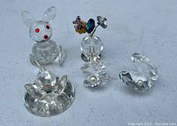 Photo showing clear Swarovski crystal bunny rabbit with red eyes, flower with colorful crystal petals on clear round base, and three faceted crystal clamshells, two of which have small pearl details.