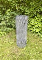 Full view of chicken wire spool standing upright on grass with leafy green plants and bushes in background.