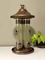 Front view of the bird feeder showing two clear plastic tubes with feeding ports and metal decorative band under a metal conical top, next to a house plant.