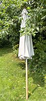 Closed beige fabric patio umbrella with wooden frame and surrounded by greenery