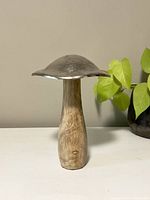 Front view of the decorative mushroom showing the wooden base and metal cap against a neutral background with a leafy plant.