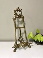 Front view of vintage brass easel with intricate floral and scroll designs, showing full ornate shape and size comparison with nearby plant.