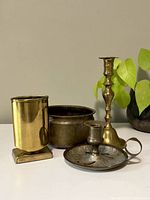 Photo showing all four brass items together on a surface with a plant in the background: two brass candlesticks, one pot, and one pen holder.