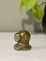 Side angled view of small brass deep sea diver bust on a plain surface with a green plant blurred in background