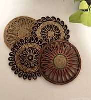 Top view of all four circular wicker trivets arranged overlapping on a white surface with a plant partially visible in the background. Shows varied woven patterns and black and red accent colors.