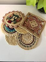 Top view showing full set of six wicker trivets stacked to display various colorful woven and embroidered patterns, round and square shapes visible.