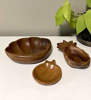 Front and slightly downward view of three wooden bowls on white surface: large seashell-shaped, medium pineapple-shaped, small pear-shaped.