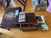 View of antique wooden sewing box, tapestry foot stool, wooden wall hook rack, and plastic container with sewing notions all grouped together on floor.