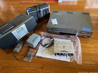 Overview of all items showing Samsung DVD-VCR unit, Sony CD player boom box with remote, cassette recorder with broken handle, and miscellaneous accessories and manuals spread on wooden floor.
