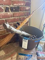 Photo showing tin bucket with fire log, large pinecone, fern broom leaning on bucket, and metal firetools inside bucket, placed near brick fireplace.