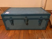 Top and front view of blue metal footlocker sitting on wooden floor. Shows latch and clasp closure, metal reinforcements on corners.