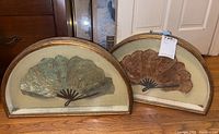 Two vintage crescent-shaped fans framed behind glass, placed on wooden surface near furniture.