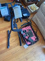 Black metal guitar rack with padded arms positioned upright with a tag reading 'Guitar Rack + Computers'. Electronics accessories including a numeric keypad, chargers, and cables placed on the floor next to it.