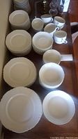 Wide view of entire dinnerware collection showing stacked white plates, bowls, coffee cups, and saucers on a wooden surface.