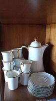 Full set view showing stacked cups, saucers, and the tall tea/coffee pot with lid, displaying the hand-painted gold rims and handles on white porcelain.