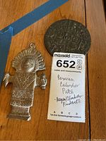 Photo of the round metal Peruvian calendar patch and the standing figure metal pendant with label showing lot number and description.