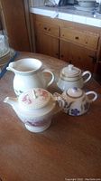 Set of 4 items on wooden table: 3 teapots and 1 water pitcher. View includes table setting and kitchen background.