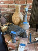 Photo showing the tin turtle figurine on a brick surface, next to the woven jar and tall vase.